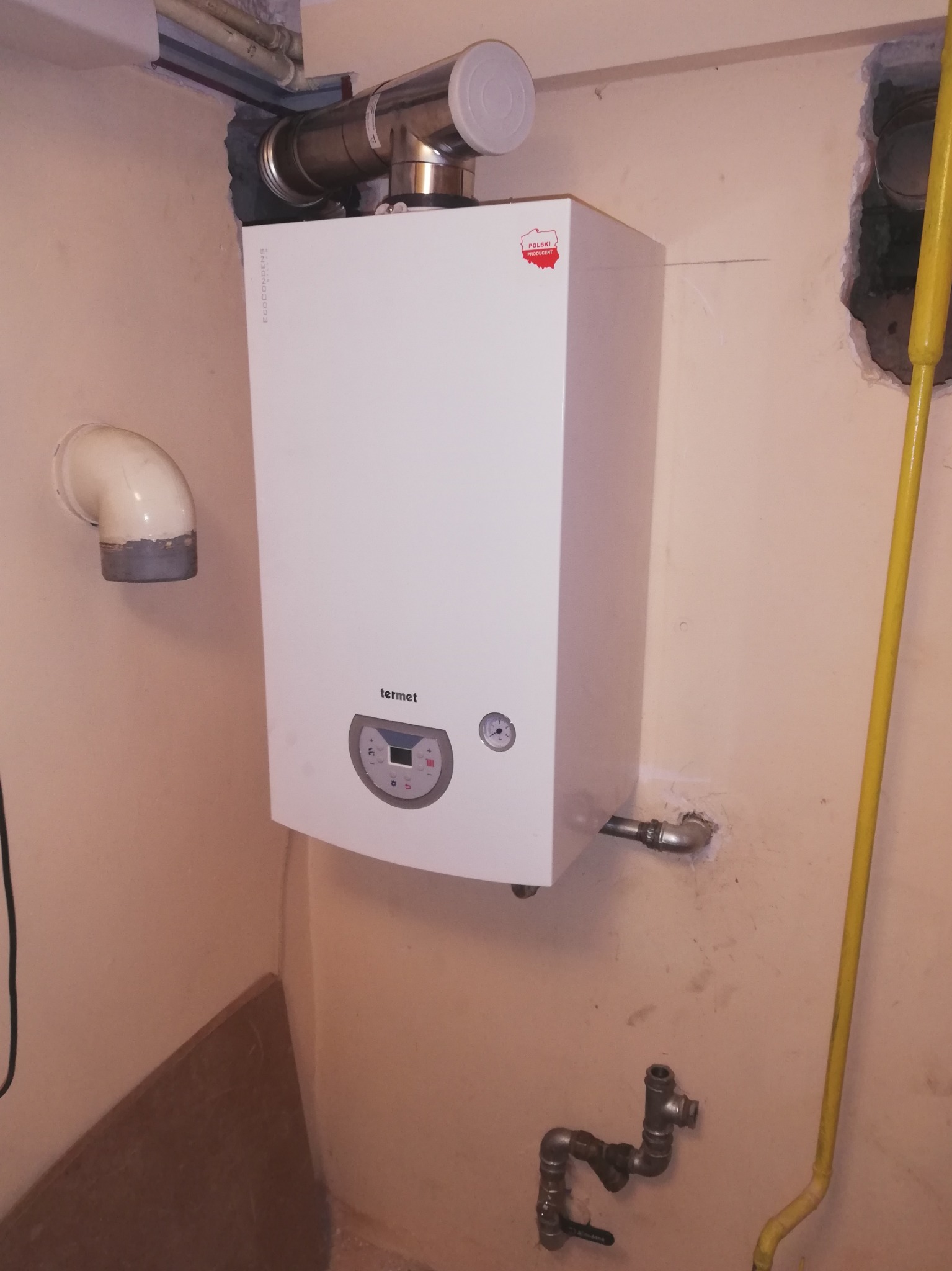 Biały piec gazowy marki Termet, zamontowany na beżowej ścianie z widocznymi rurami i instalacją hydrauliczną, z naklejką 'Polski Producent'.
