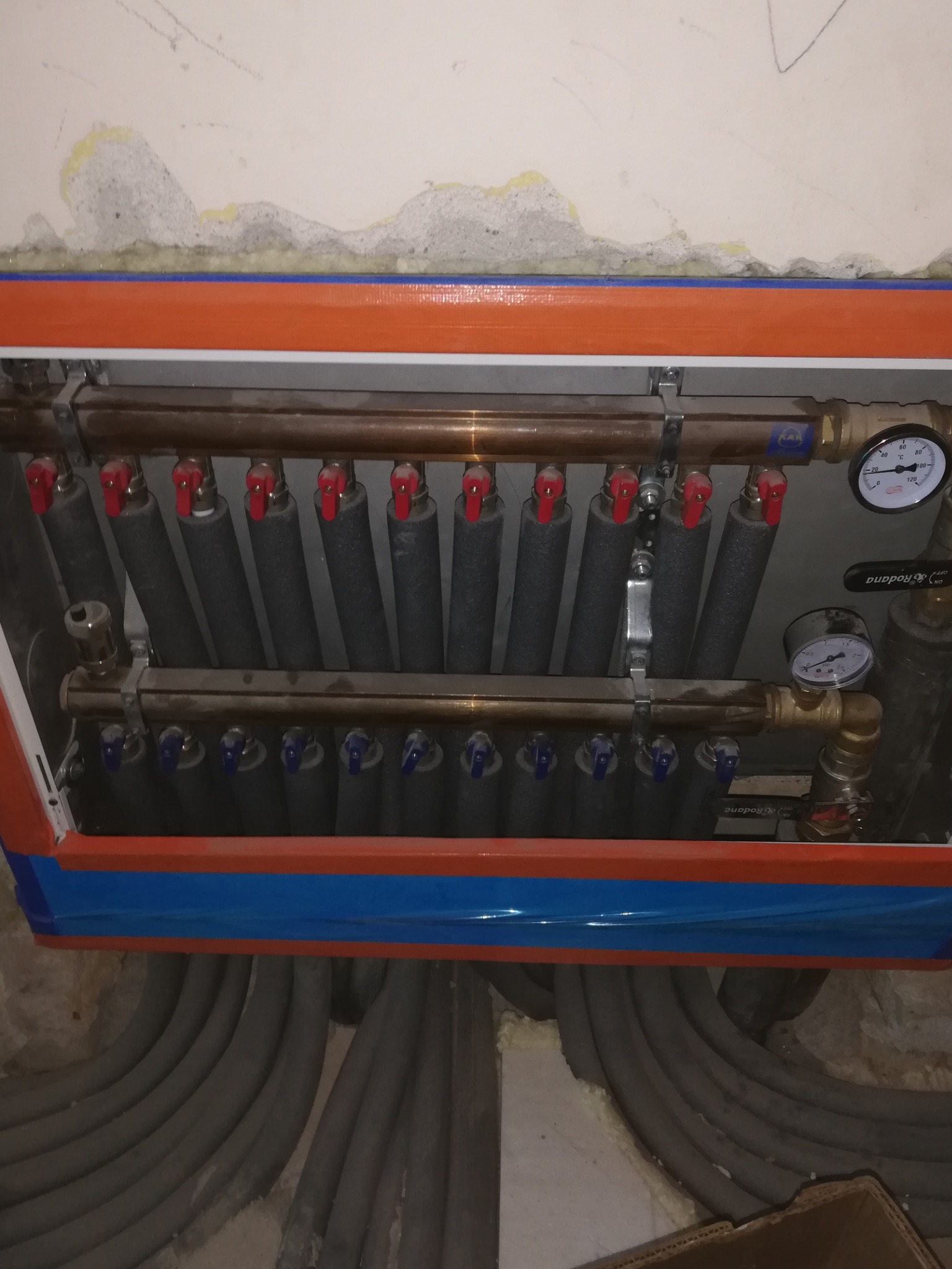 Rozdzielacz ogrzewania podłogowego z zaworami, manometrami i izolowanymi rurami, zamontowany w obudowie z pomarańczowo-niebieskim wykończeniem, z widocznymi szarymi rurami instalacji. Ściana w tle...