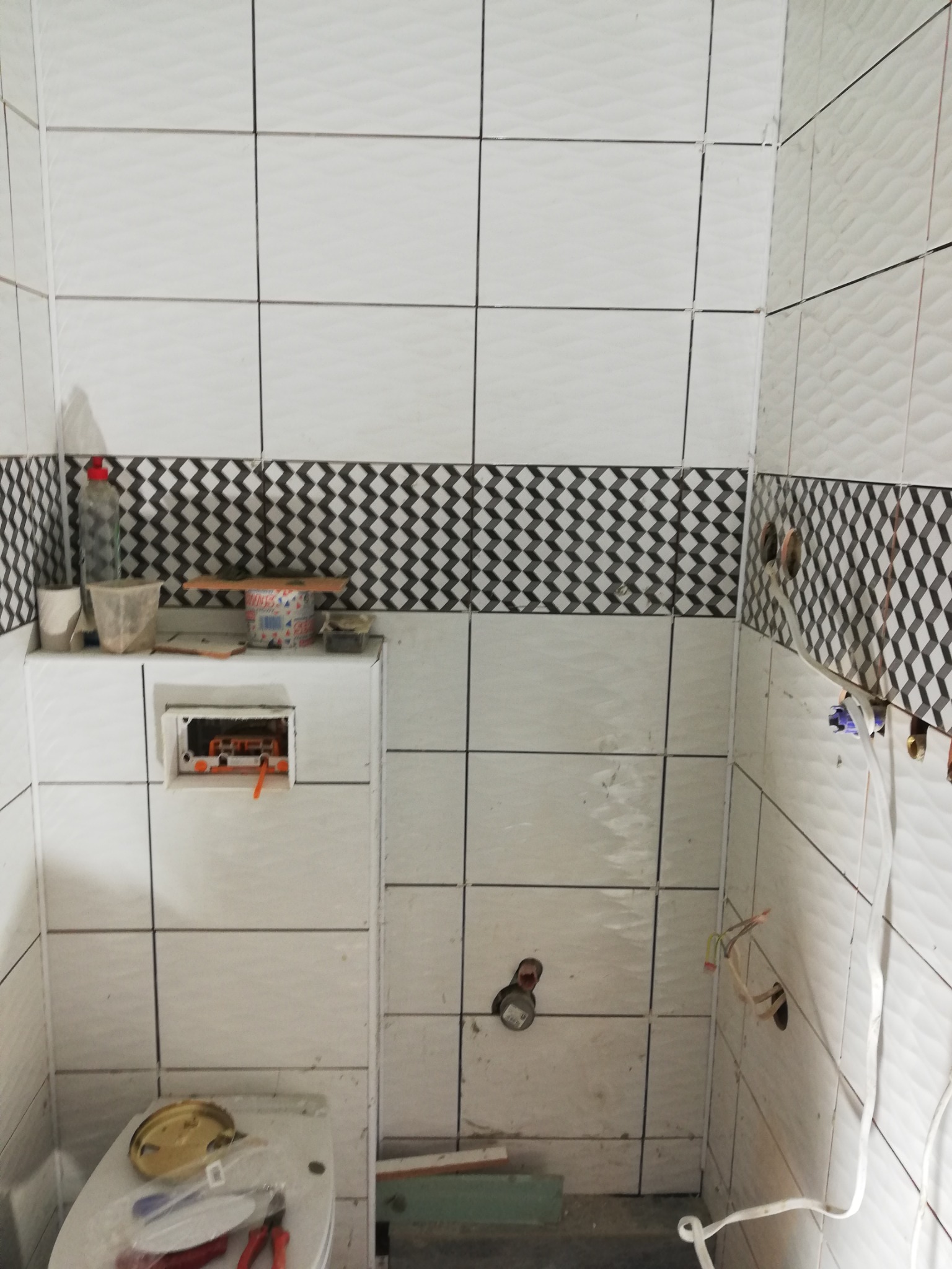 Łazienka w trakcie wykańczania: białe płytki z dekoracyjnym pasem w zygzaki, widoczne instalacje elektryczne i hydrauliczne, narzędzia i materiały budowlane na toalecie.