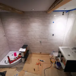 Łazienka w trakcie remontu: widoczne płytki na ścianach, częściowo zamontowana umywalka z szafką, kabina prysznicowa w rogu, narzędzia i materiały budowlane rozłożone na podłodze zabezpieczonej...