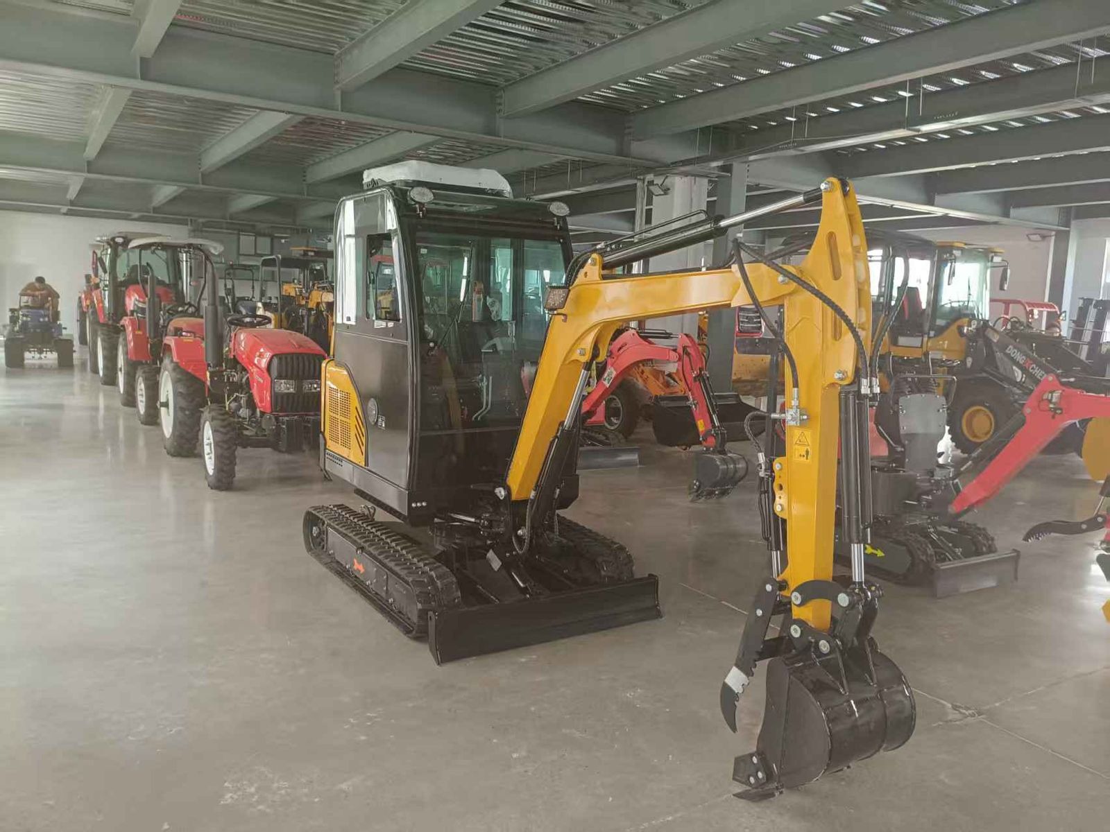Żółto-czarna mini koparka gąsienicowa z chwytakiem na tle rzędu traktorów w hali. Sprzęt budowlany w magazynie, widok z boku, oświetlenie górne.
