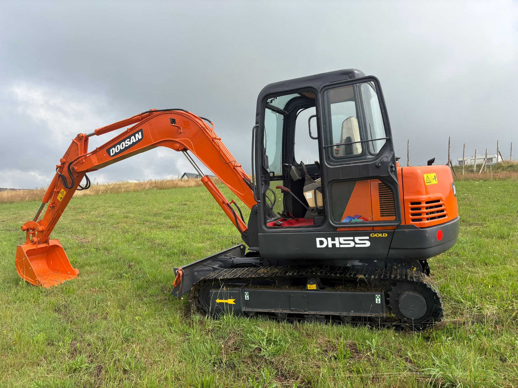 Pomarańczowa mini koparka Doosan DHS55 Gold na zielonej trawie pod pochmurnym niebem. Widoczny lemiesz i kabina operatora. Maszyna gotowa do pracy.