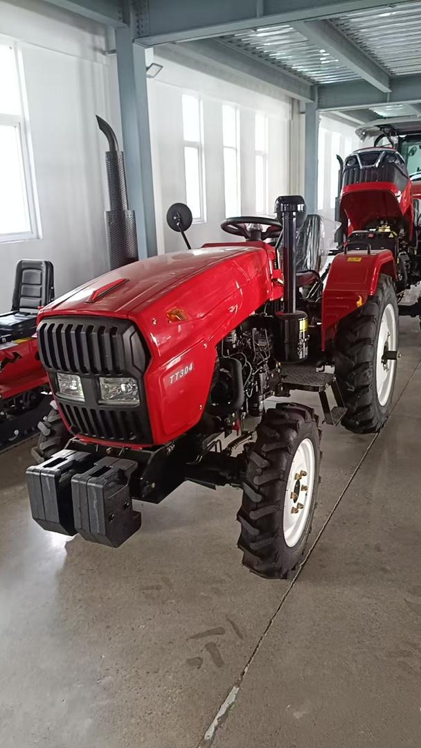 Czerwony traktor TT304 z czarną atrapą chłodnicy i obciążnikami na tle hali z oknami. Widoczny fragment drugiego traktora.