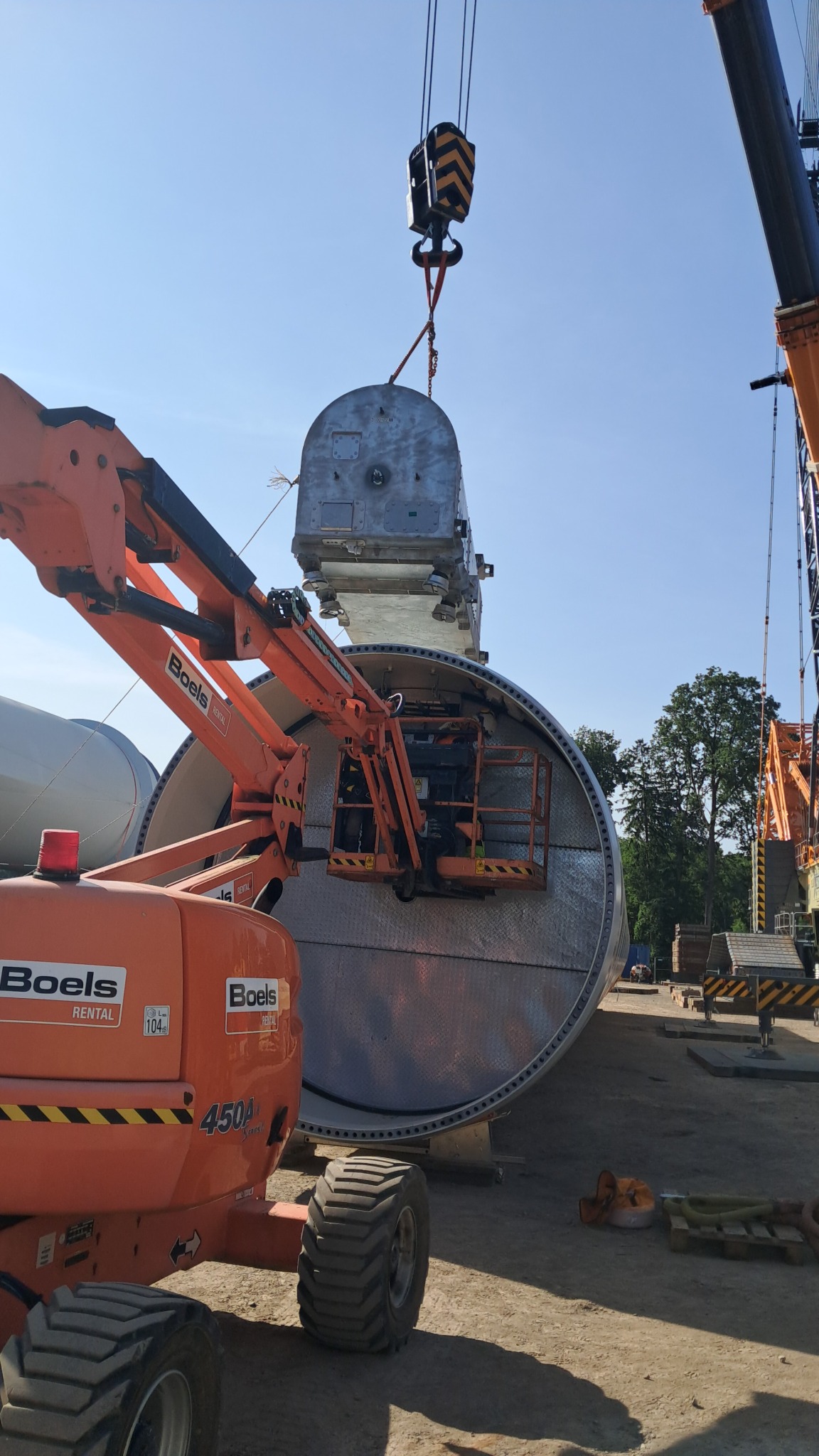 Montaż turbiny wiatrowej: dźwig podnosi element do gondoli, widoczny podnośnik koszowy i fragmenty innych elementów turbiny. Słoneczny dzień, plac budowy.