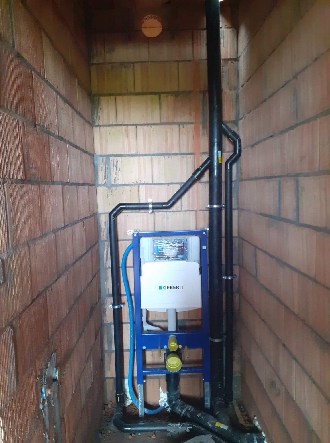 Stelaż podtynkowy Geberit Duofix zamontowany w surowej ścianie z cegły, z widocznymi czarnymi rurami kanalizacyjnymi i niebieską rurą doprowadzającą wodę.