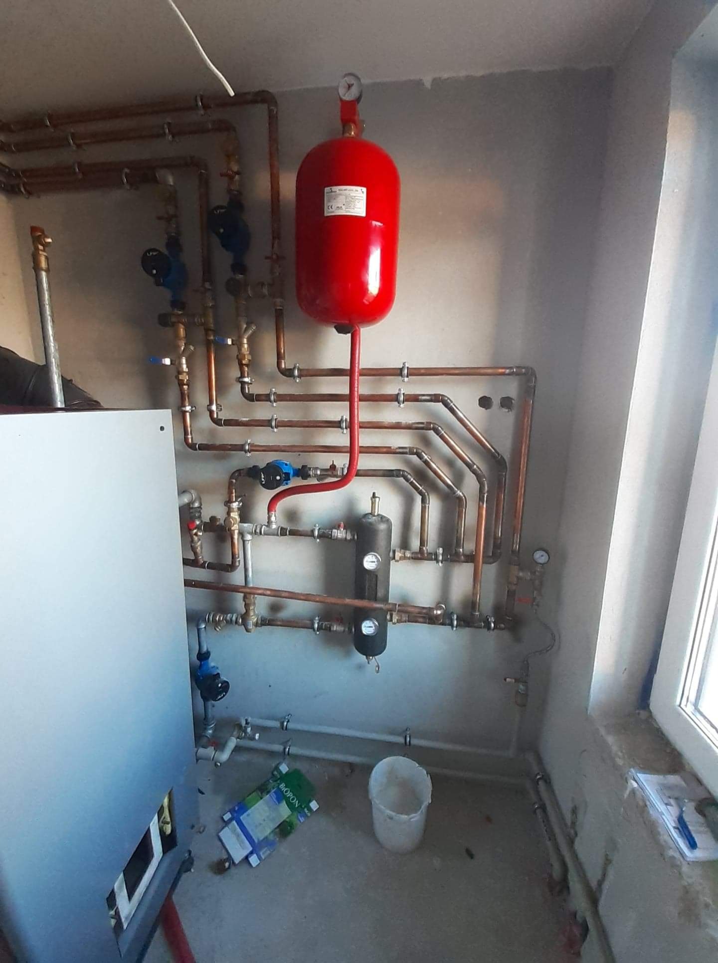 Świeżo zainstalowana instalacja centralnego ogrzewania z czerwonym zbiornikiem wyrównawczym, miedzianymi rurami i czarnym naczyniem przeponowym, widoczna na tle surowej ściany i okna w stanie...