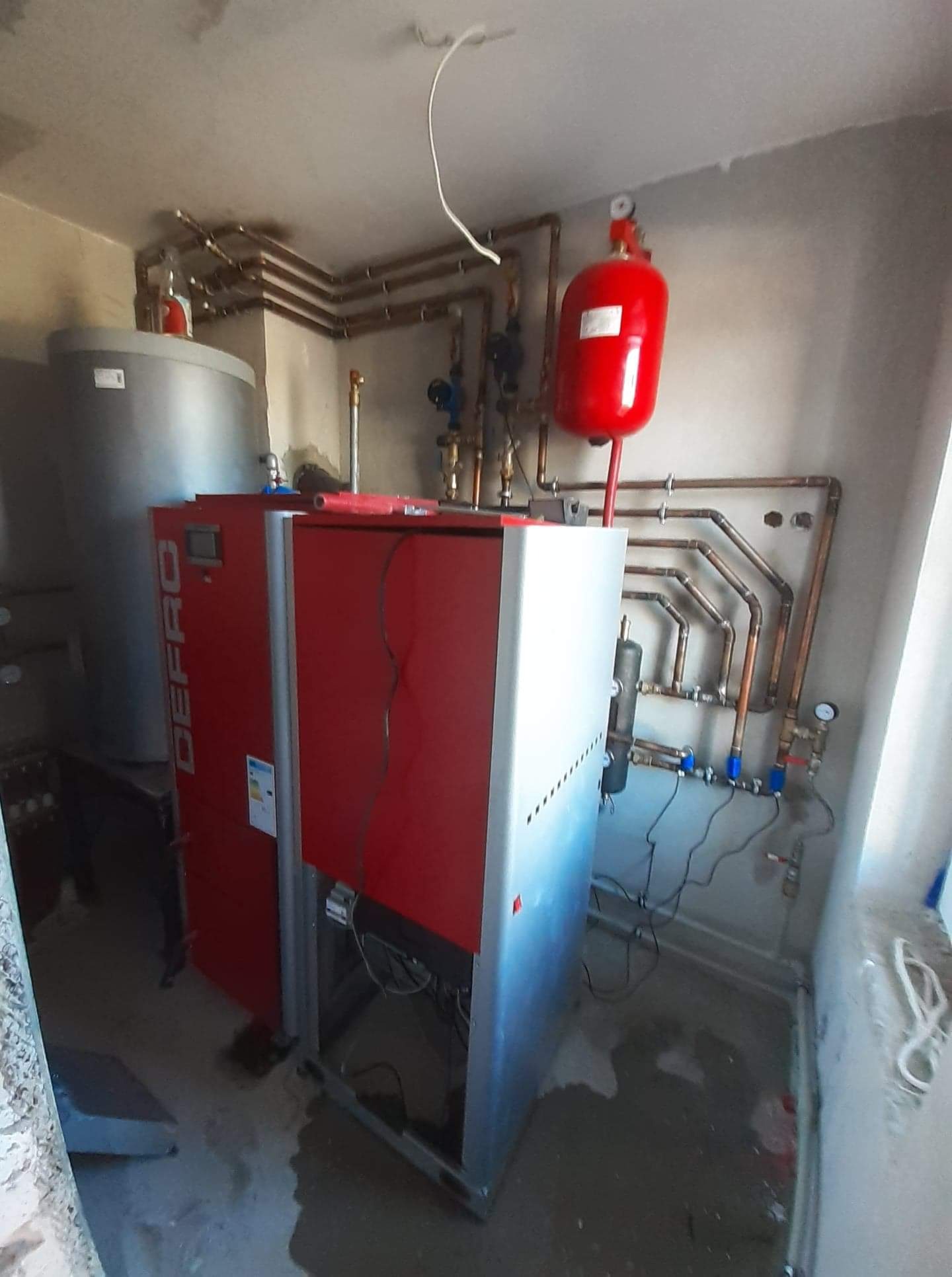 Nowo zainstalowany, czerwono-srebrny piec grzewczy DEFRO z podłączonymi miedzianymi rurami i czerwonym zbiornikiem wyrównawczym w surowym pomieszczeniu.
