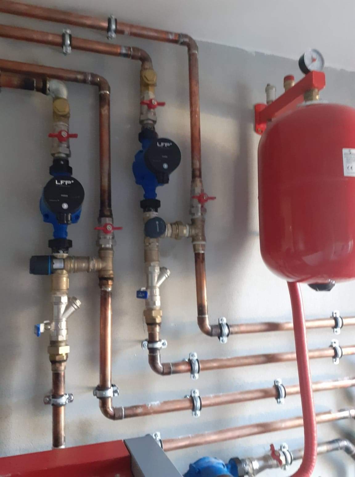 Instalacja centralnego ogrzewania z miedzianymi rurami, zaworami z czerwonymi pokrętłami, niebieskimi pompami obiegowymi LFP i czerwonym zbiornikiem wyrównawczym z manometrem, zamontowana na szarej...