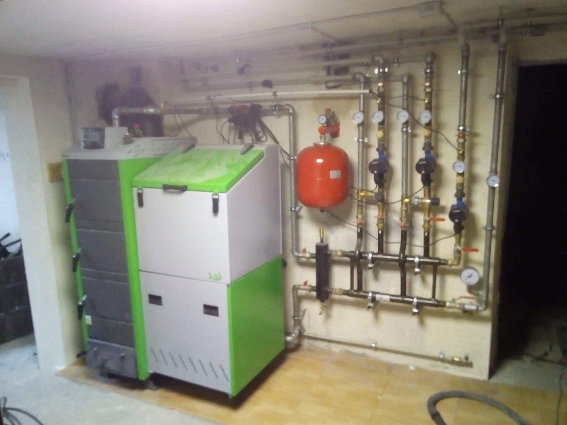 Kotłownia z piecem na paliwo stałe z zielonymi akcentami i instalacją hydrauliczną z manometrami, widok ogólny na tle ściany z rurami.