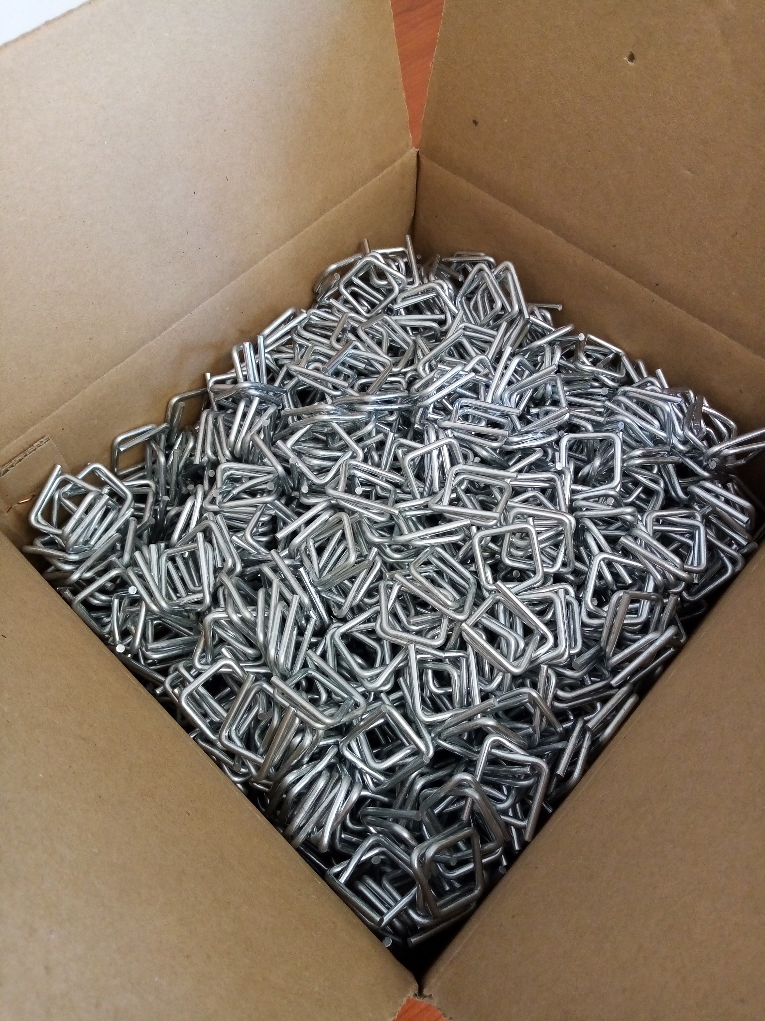 Karton wypełniony metalowymi, srebrnymi klamrami w kształcie litery U, przeznaczonymi do mocowania materiałów.