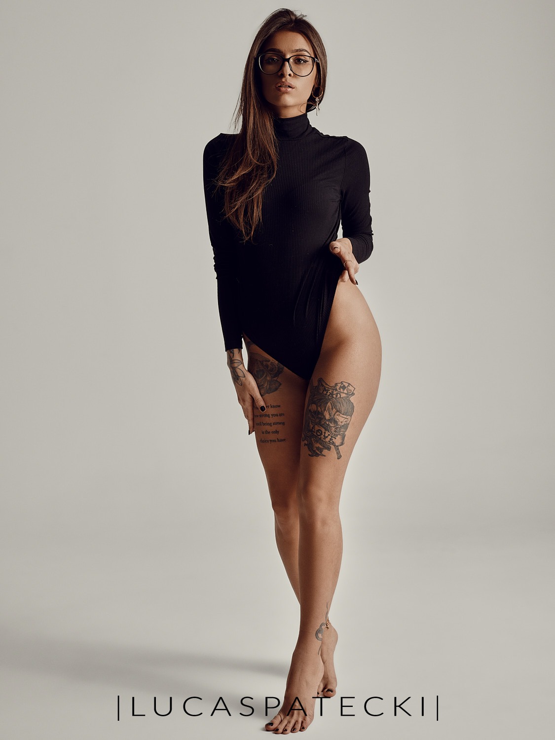 Kobieta w czarnym body, okularach i z tatuażami na udach, pozująca na jasnym tle w studio fotograficznym.