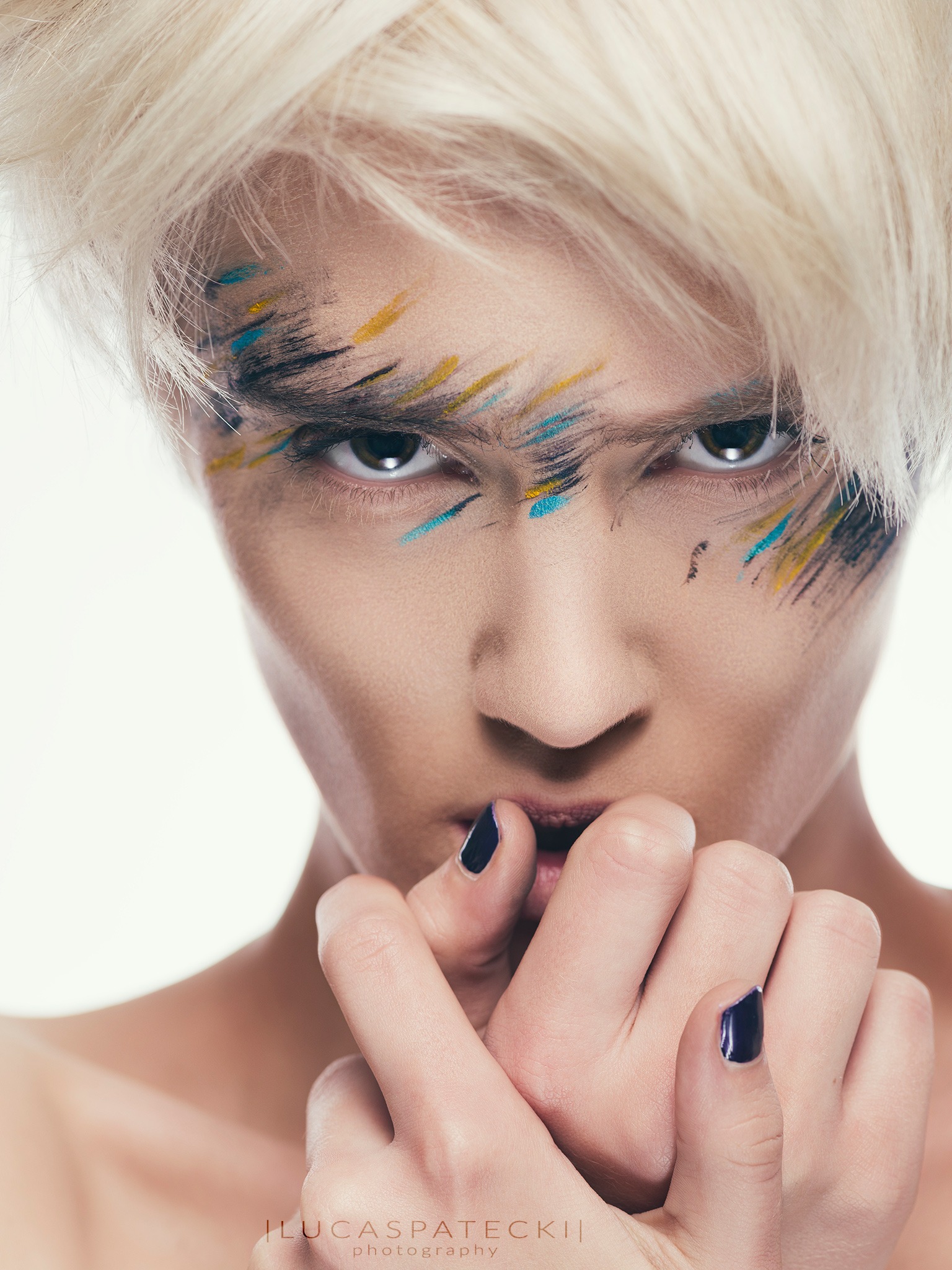 Portret kobiety z krótkimi blond włosami i artystycznym makijażem: czarne, niebieskie i żółte pociągnięcia pędzlem wokół oczu, usta lekko otwarte, dłonie z ciemnym lakierem na paznokciach przy ustach.