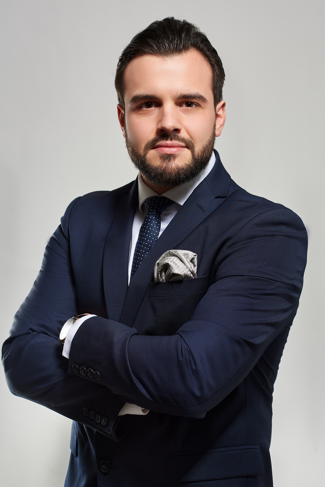 Portret mężczyzny w granatowym garniturze w kratę, z założonymi rękami, widoczny zegarek i poszetka w kieszonce.