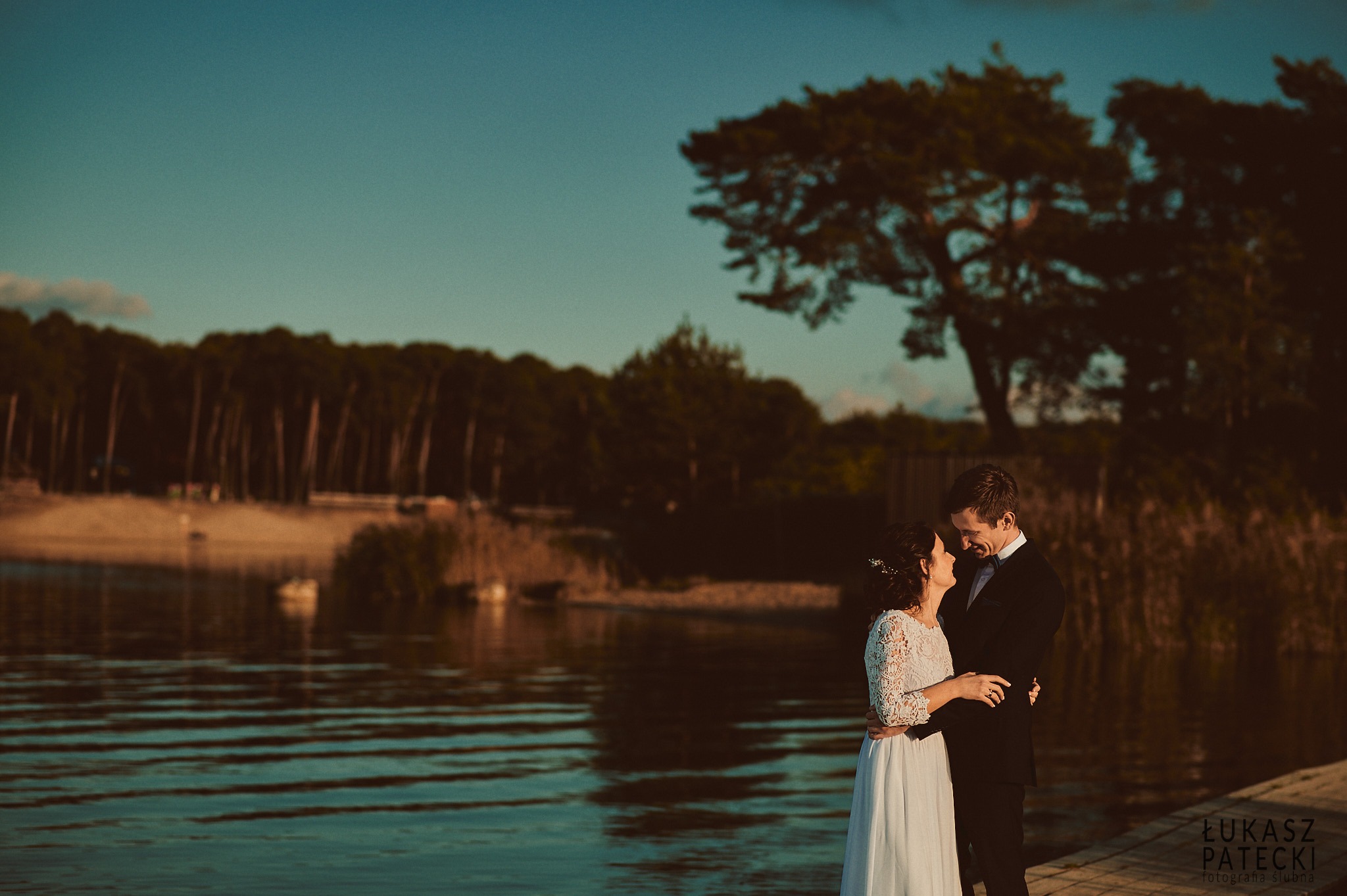 Para młoda w objęciach na tle jeziora i drzew podczas zachodu słońca; kobieta w sukni z koronkowymi rękawami, mężczyzna w czarnym garniturze i muszce.