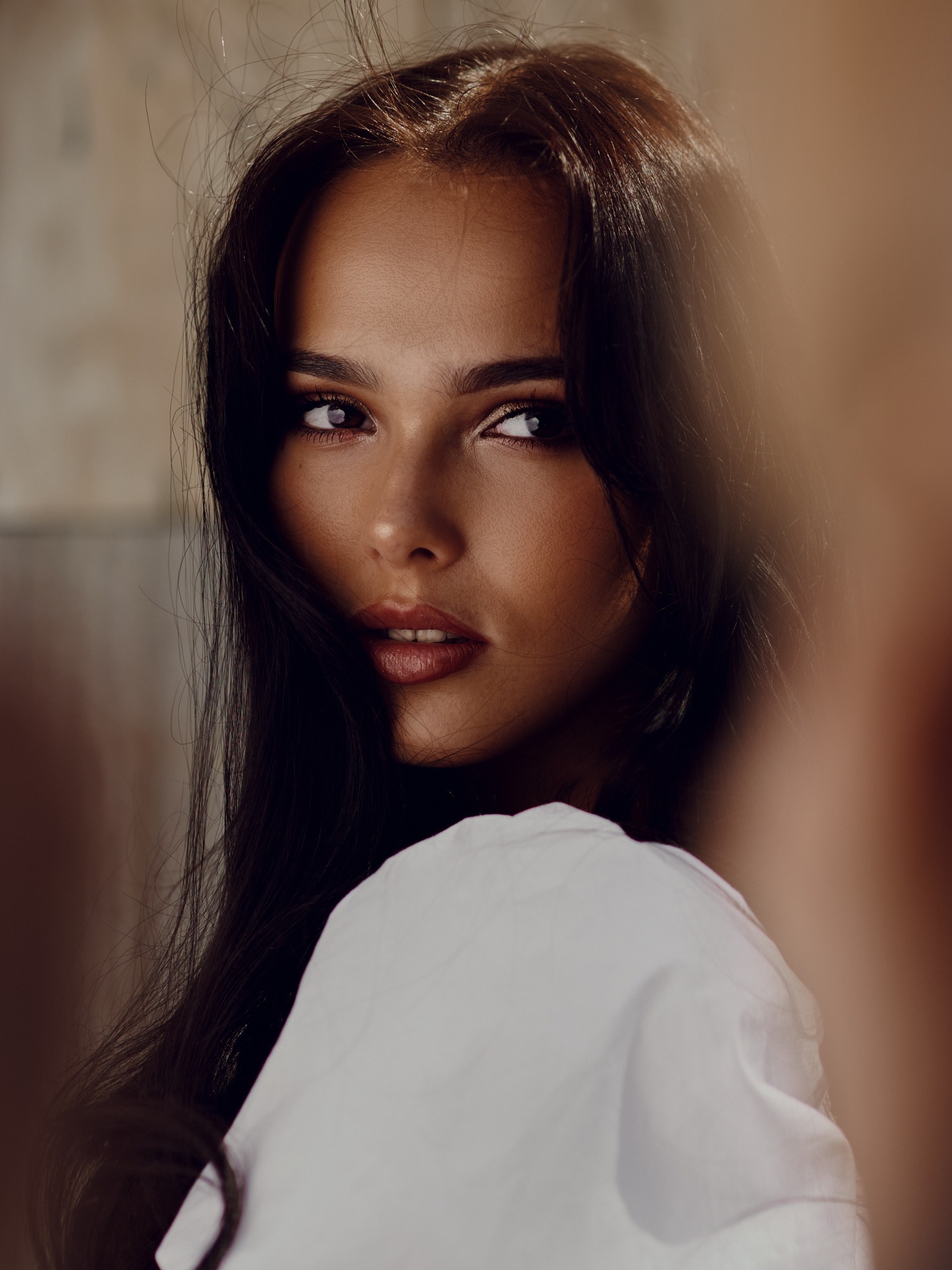 Portret kobiety z ciemnymi włosami, spoglądającej przez ramię, z delikatnym makijażem i białej koszuli na rozmytym tle.