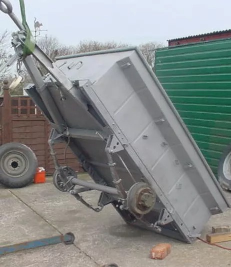 Przyczepa rolnicza podnoszona na haku, świeżo pokryta szarą farbą, widoczne detale podwozia i koła, tło z zielonym kontenerem i ogrodzeniem.