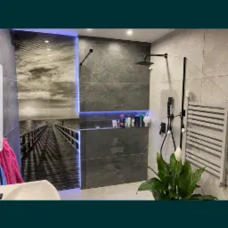 Nowoczesna łazienka z szarymi płytkami, deszczownicą w kolorze czarnym, podświetlaną wnęką z kosmetykami i grafiką na ścianie przedstawiającą molo.