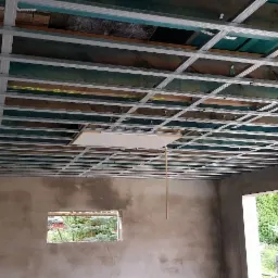 Metalowa konstrukcja sufitowa podwieszana z widocznymi elementami mocującymi, surowy stan ścian w pomieszczeniu z oknem na zewnątrz.