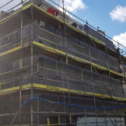 Budynek w trakcie ocieplania, pokryty czarną izolacją i otoczony rusztowaniem z żółtymi elementami, widok na budynek mieszkalny w tle, błękitne niebo z chmurami.