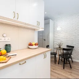 Jasna kuchnia z białymi szafkami, drewnianym blatem, ścianą z białej cegły, czarnymi krzesłami i drewnianą podłogą, z warzywami i owocami na blacie.