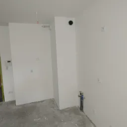 Wnętrze w stanie deweloperskim z gładkimi, pomalowanymi na biało ścianami, surową posadzką w odcieniach szarości oraz wystającymi instalacjami hydraulicznymi i elektrycznymi.
