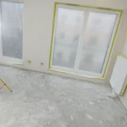 Przygotowanie do malowania w Krakowie: Pok&oacute;j z zabezpieczonymi oknami folią i taśmą malarską, surowy beton na podłodze z plamami farby, widoczna wisząca żar&oacute;wka.