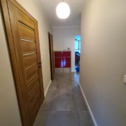 Korytarz z szarymi płytkami podłogowymi, beżowymi ścianami, drewnianymi drzwiami z poziomymi przeszkleniami i lampą sufitową w kształcie kuli.