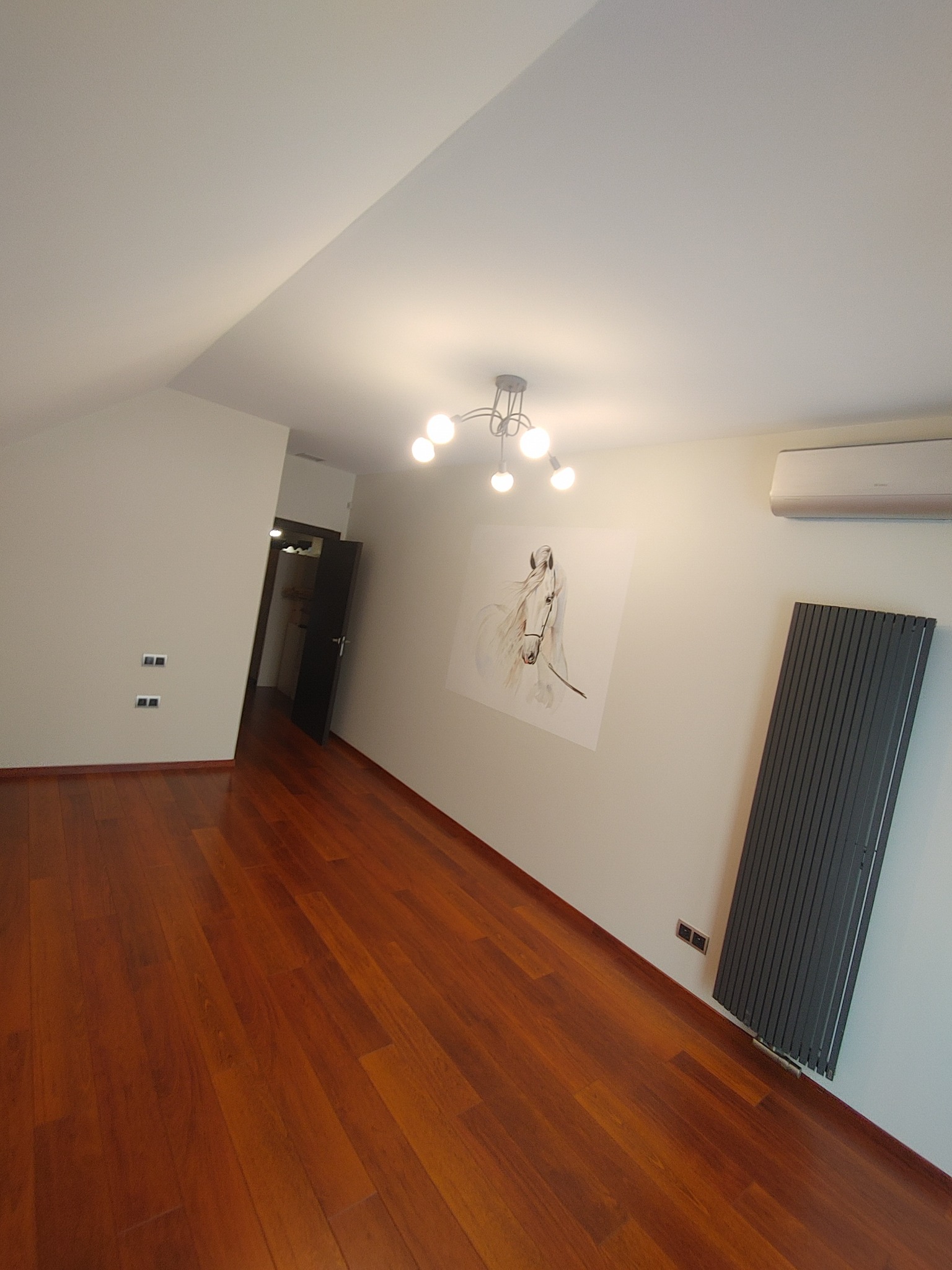 Jasne wnętrze pod skosem z parkietem w odcieniach brązu, grzejnikiem panelowym, klimatyzatorem, lampą sufitową z kilkoma żarówkami i obrazkiem konia na ścianie.