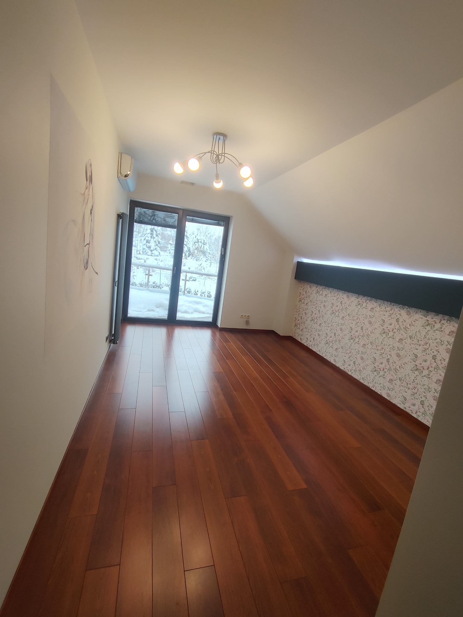 Poddasze z drewnianą podłogą, ścianą z tapetą w kwiaty i wyjściem na balkon z widokiem na zimowy krajobraz, oświetlone przez nowoczesną lampę sufitową.