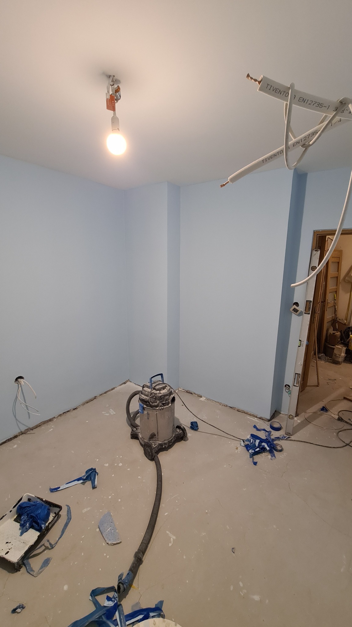 Świeżo pomalowane na błękitno ściany pokoju z widocznym odkurzaczem przemysłowym, resztkami taśmy malarskiej na podłodze i niezabezpieczonymi przewodami elektrycznymi pod sufitem, w tle widoczna...