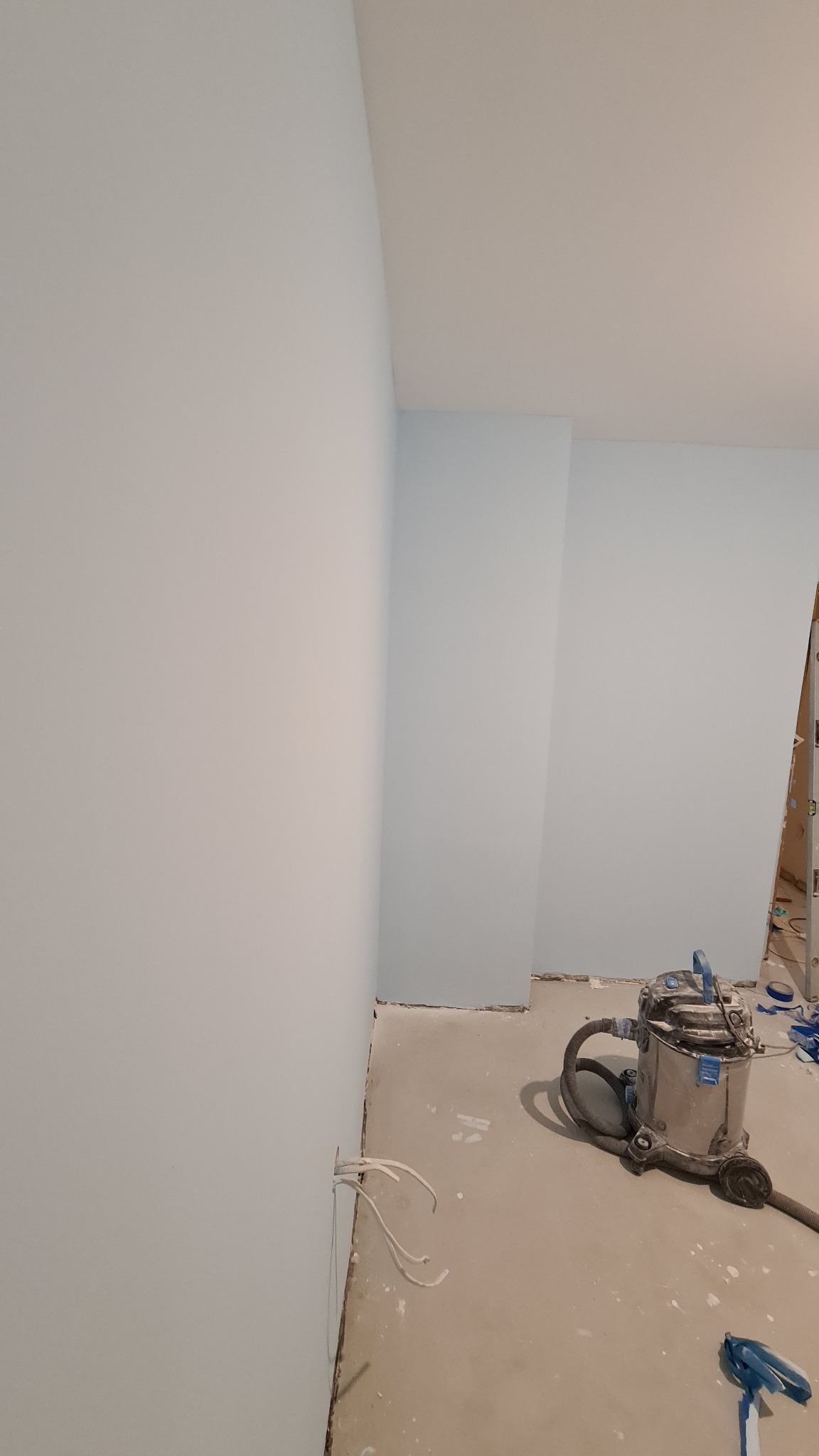 Świeżo pomalowane na błękitno ściany w pomieszczeniu, z widocznym odkurzaczem przemysłowym i wystającymi kablami ze ściany, przygotowane do dalszych prac wykończeniowych.