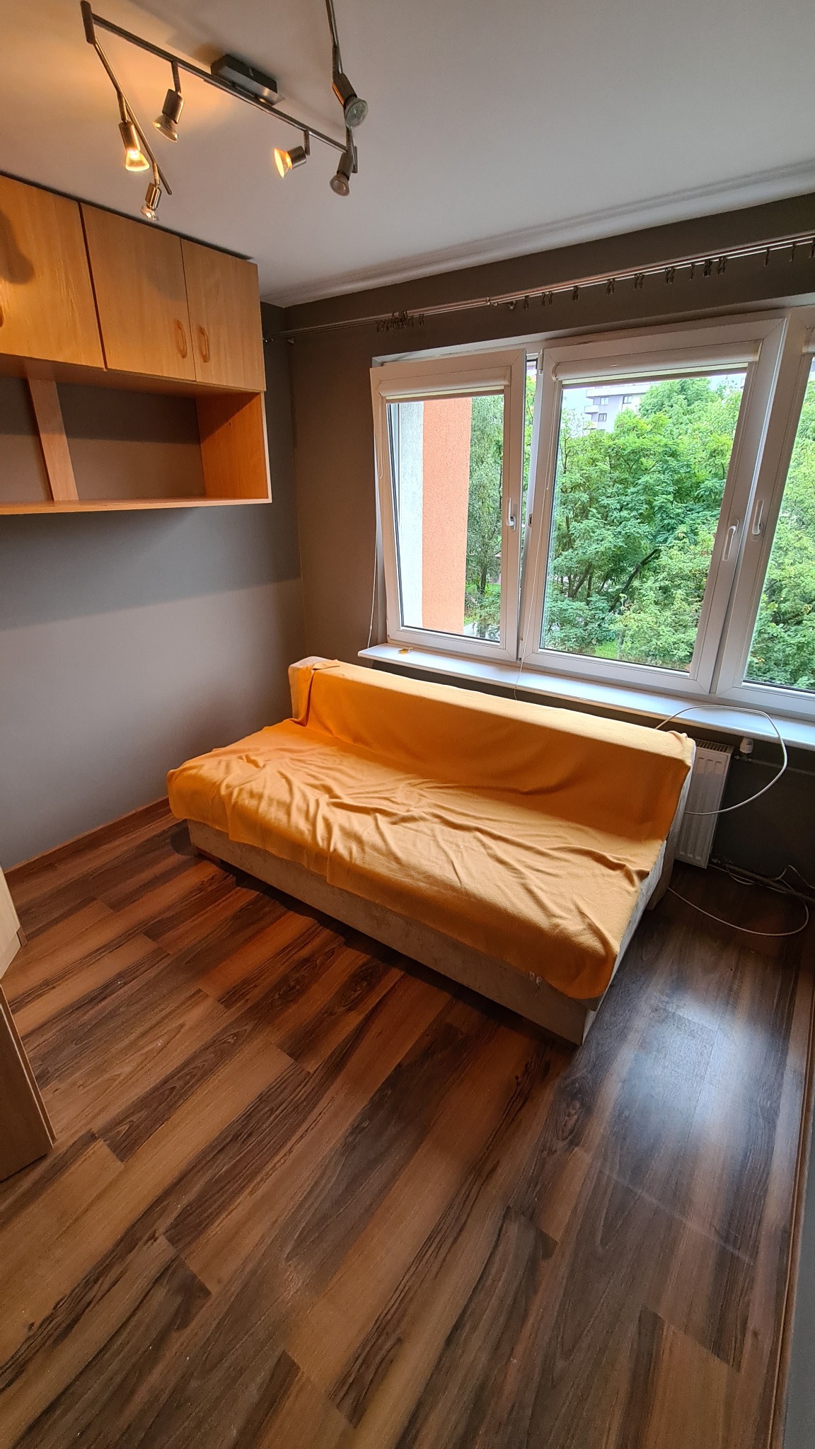 Pokój z brązowymi panelami podłogowymi, szarą ścianą i drewnianymi szafkami pod sufitem, z widokiem na zieleń za oknem, gdzie widoczna jest rozłożona sofa przykryta pomarańczową narzutą.