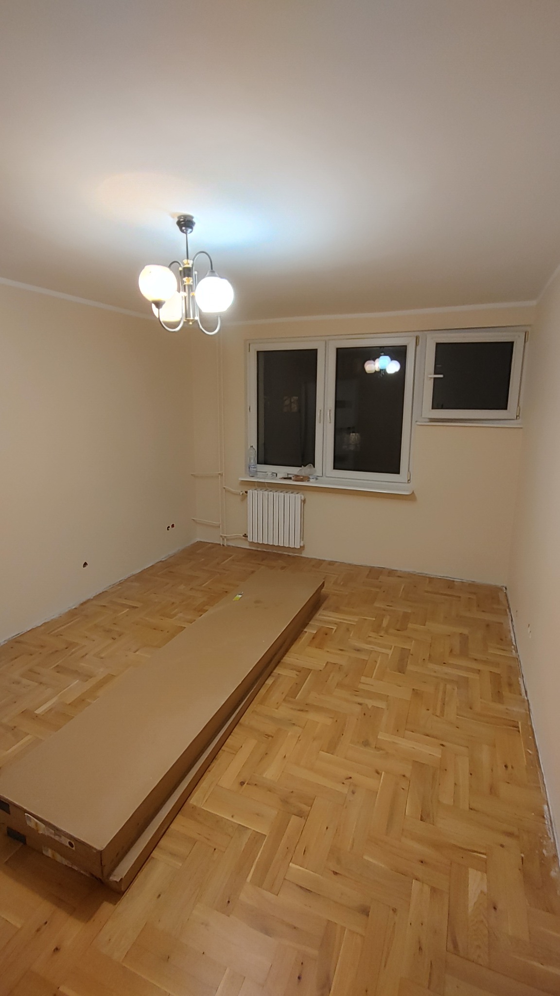 Pusty pokój z parkietem w jodełkę, przygotowany do remontu; na podłodze leżą zapakowane panele, ściany pomalowane na jasny kolor, w oknie widoczne odbicie żyrandola.