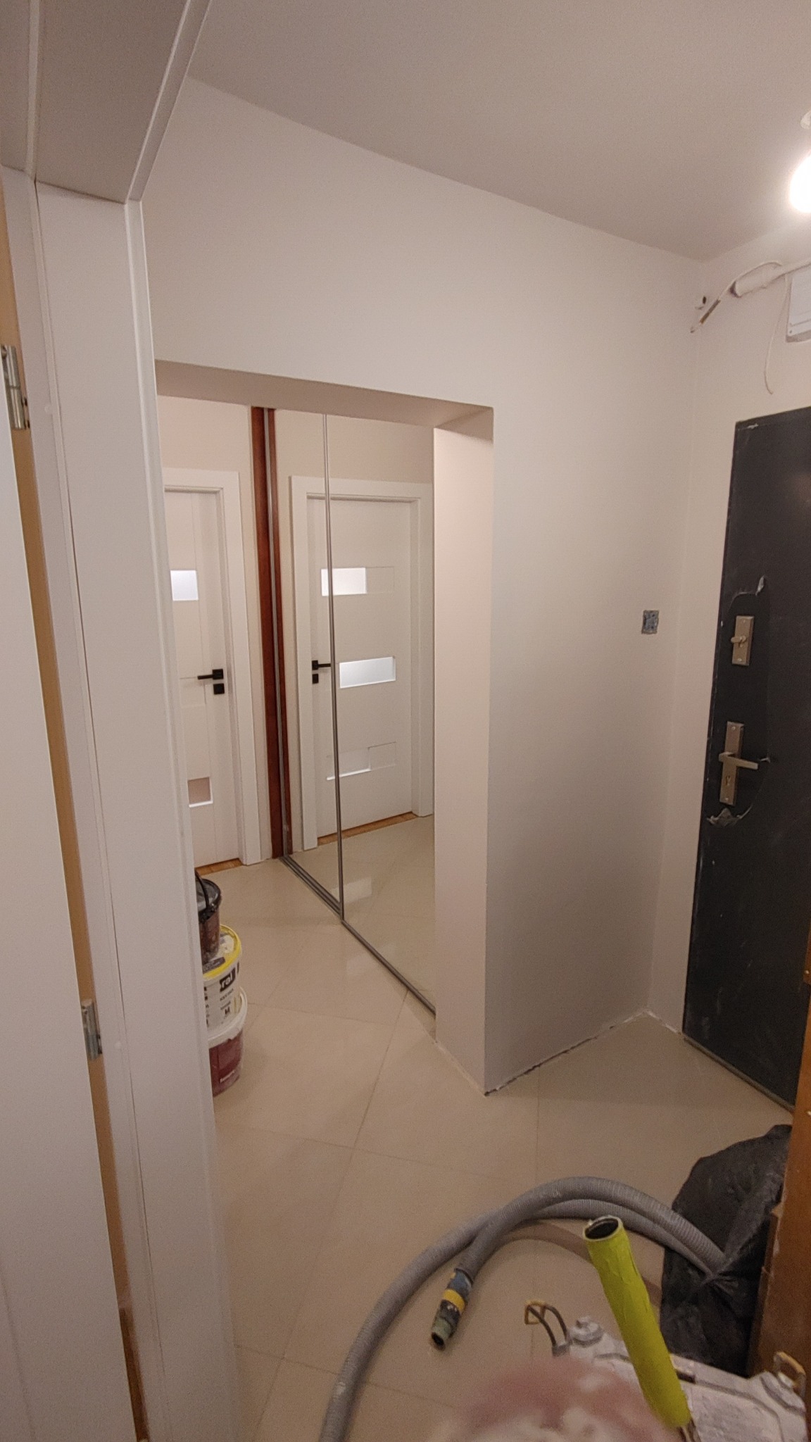 Korytarz w trakcie remontu, widoczne ślady szpachlowania na ścianach, puste wiadra po farbie i rura od odkurzacza przemysłowego na podłodze, w tle lustrzane drzwi przesuwne.
