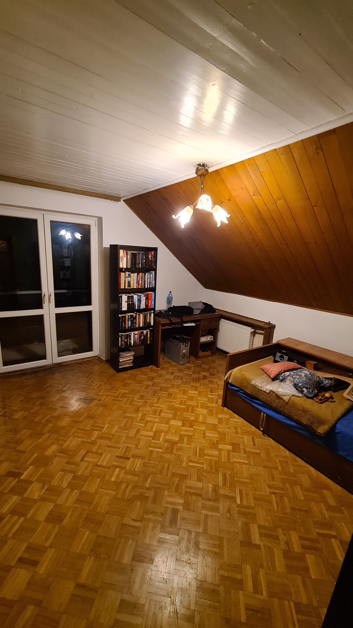 Pokój na poddaszu z drewnianą boazerią na skosie dachu, parkietem w jodełkę, regałem z książkami, biurkiem, łóżkiem i widocznym grzejnikiem.