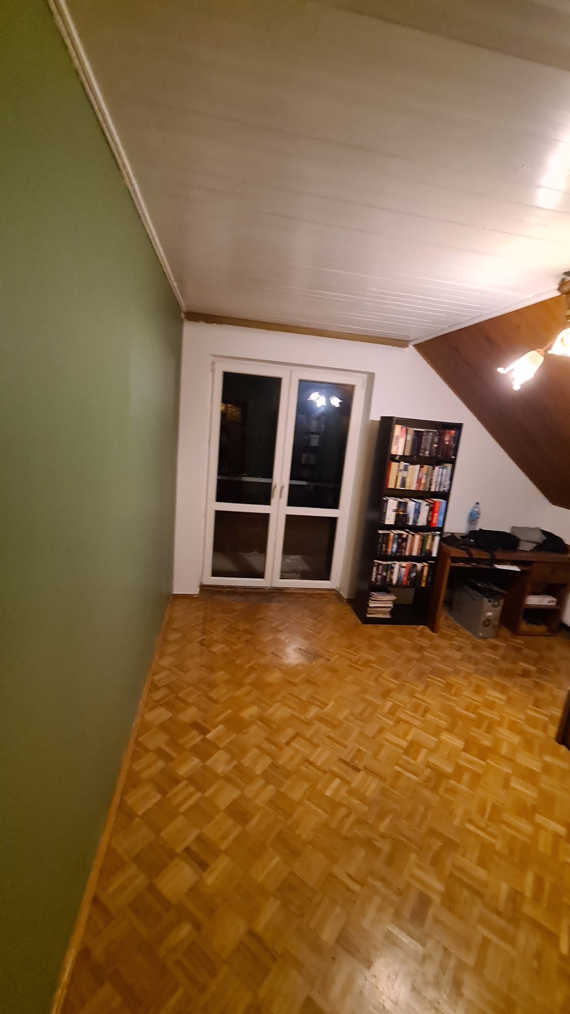 Wnętrze pokoju z parkietem ułożonym w kwadraty, zielona ściana z lewej strony, białe drzwi balkonowe na wprost, regał z książkami i biurko z prawej strony, biały sufit.