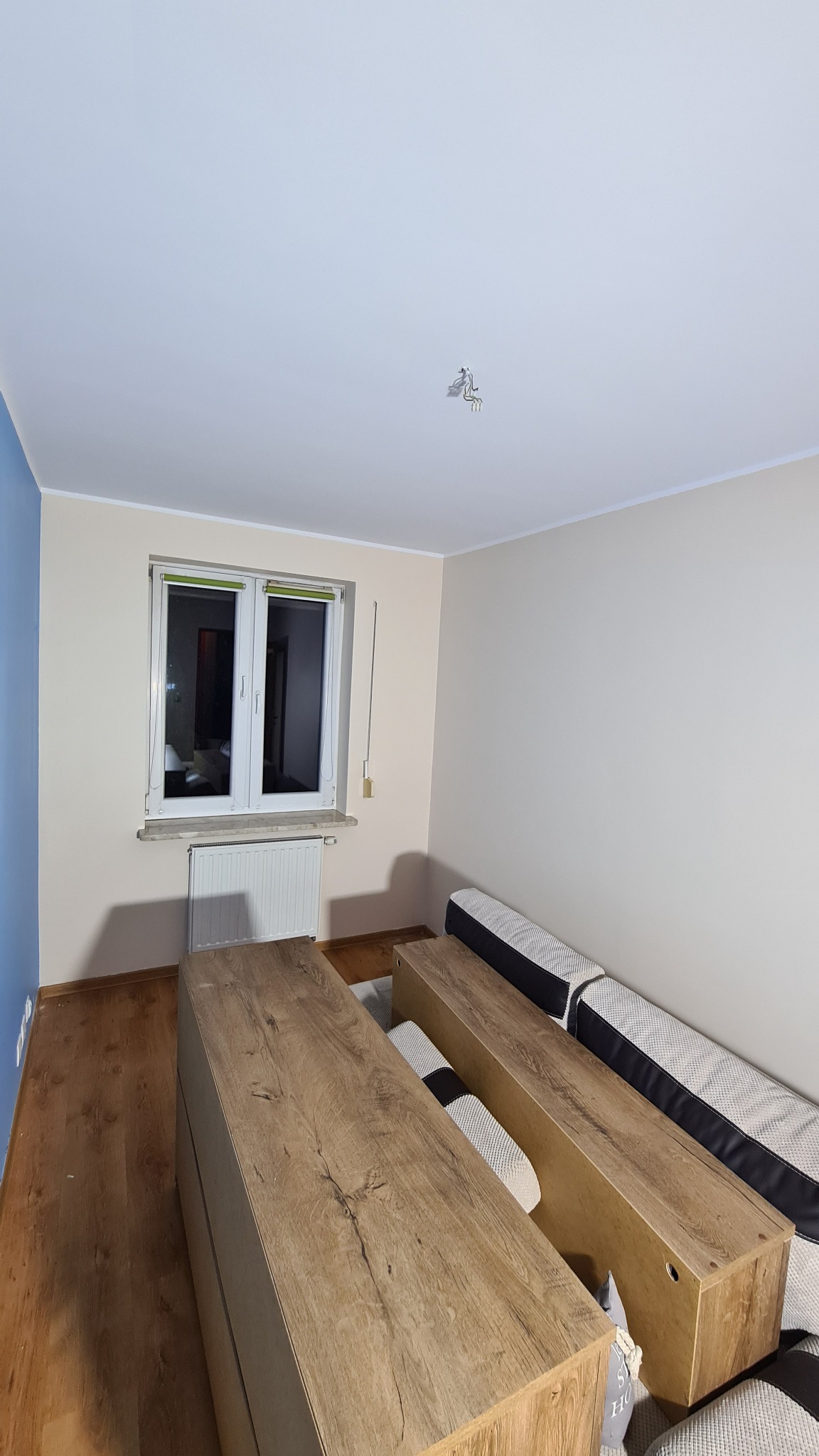 Pomalowane jasne ściany w pokoju z drewnianą podłogą, grzejnikiem pod oknem i meblami w kolorze drewna, widoczne ślady po instalacji oświetlenia na suficie.