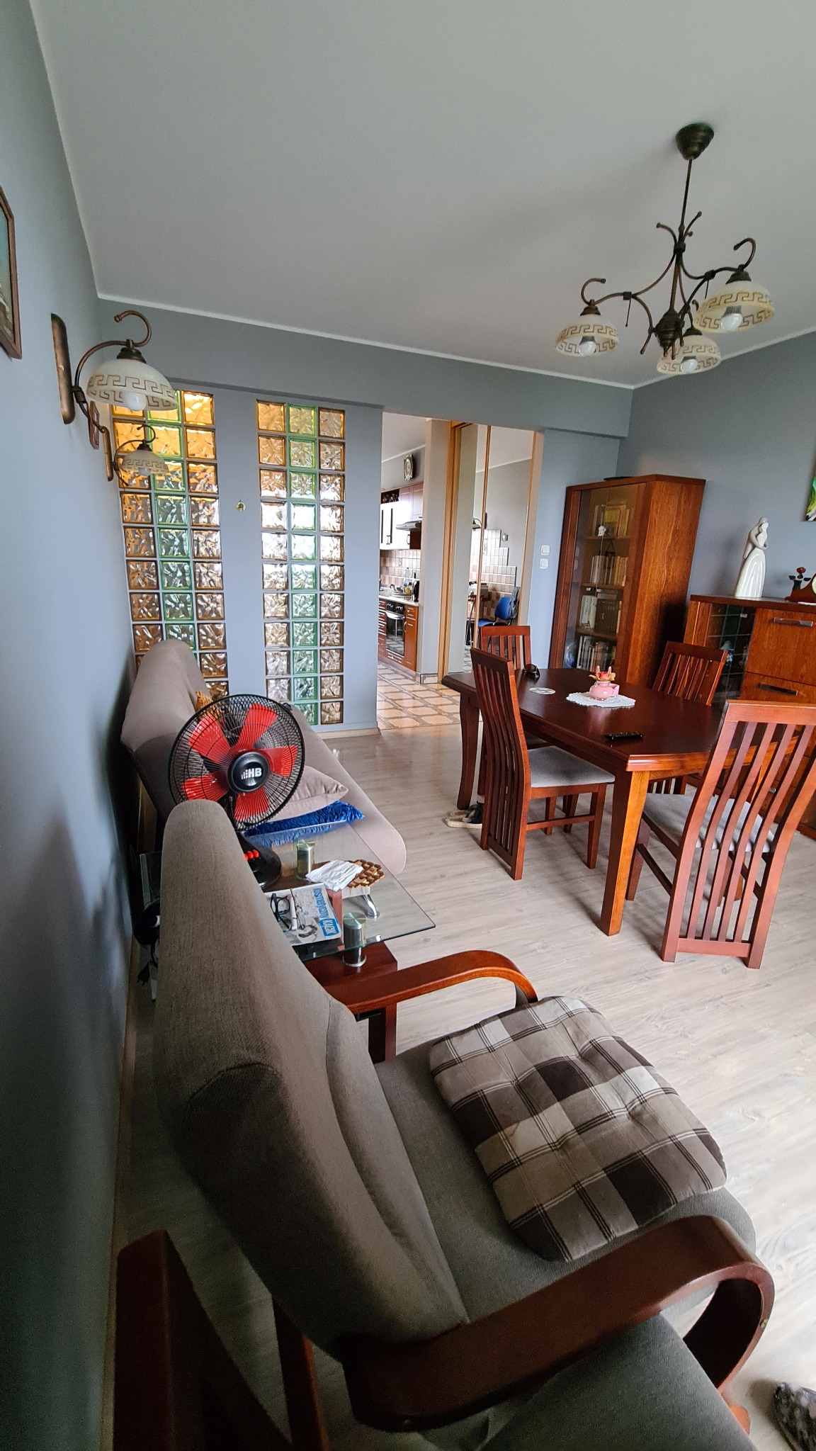 Wnętrze salonu z drewnianym stołem, krzesłami, przeszkloną witryną i ścianą z luksferów. Widoczny wentylator stojący na sofie i szary kolor ścian.