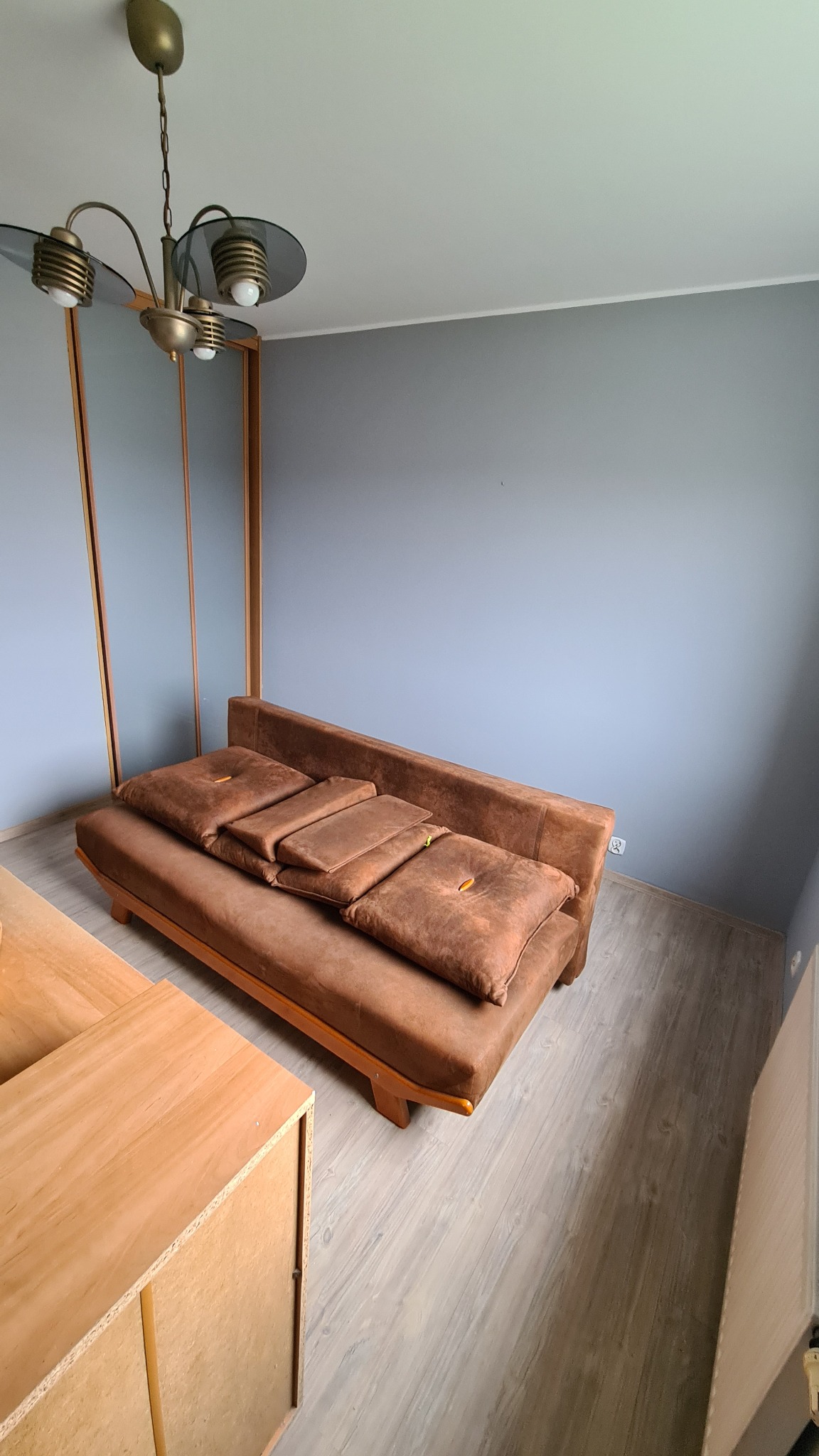 Pomalowane na szaro ściany w pokoju z brązową sofą z poduszkami, drewniana podłoga i szafka, widoczny złoty żyrandol.