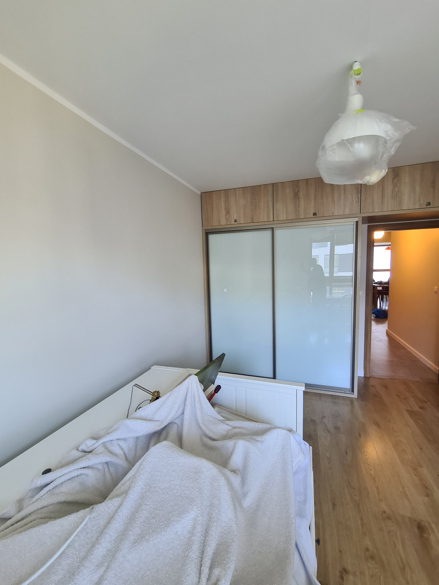 Pokój w trakcie przygotowań do malowania: ściana w jasnym odcieniu beżu, zabezpieczone meble białym materiałem, osłonięta lampa wisząca folią, widoczna część szafy przesuwnej i fragment podłogi...