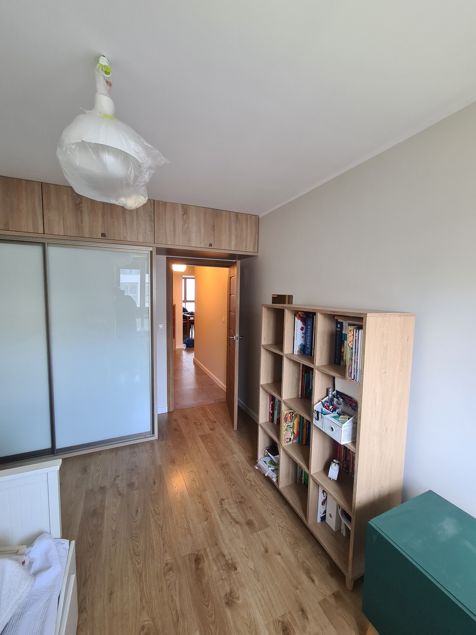 Pokój po remoncie z drewnianą podłogą, zabudowaną szafą przesuwną, regałem z książkami i lampą w folii ochronnej, widok na otwarte drzwi do dalszej części mieszkania.