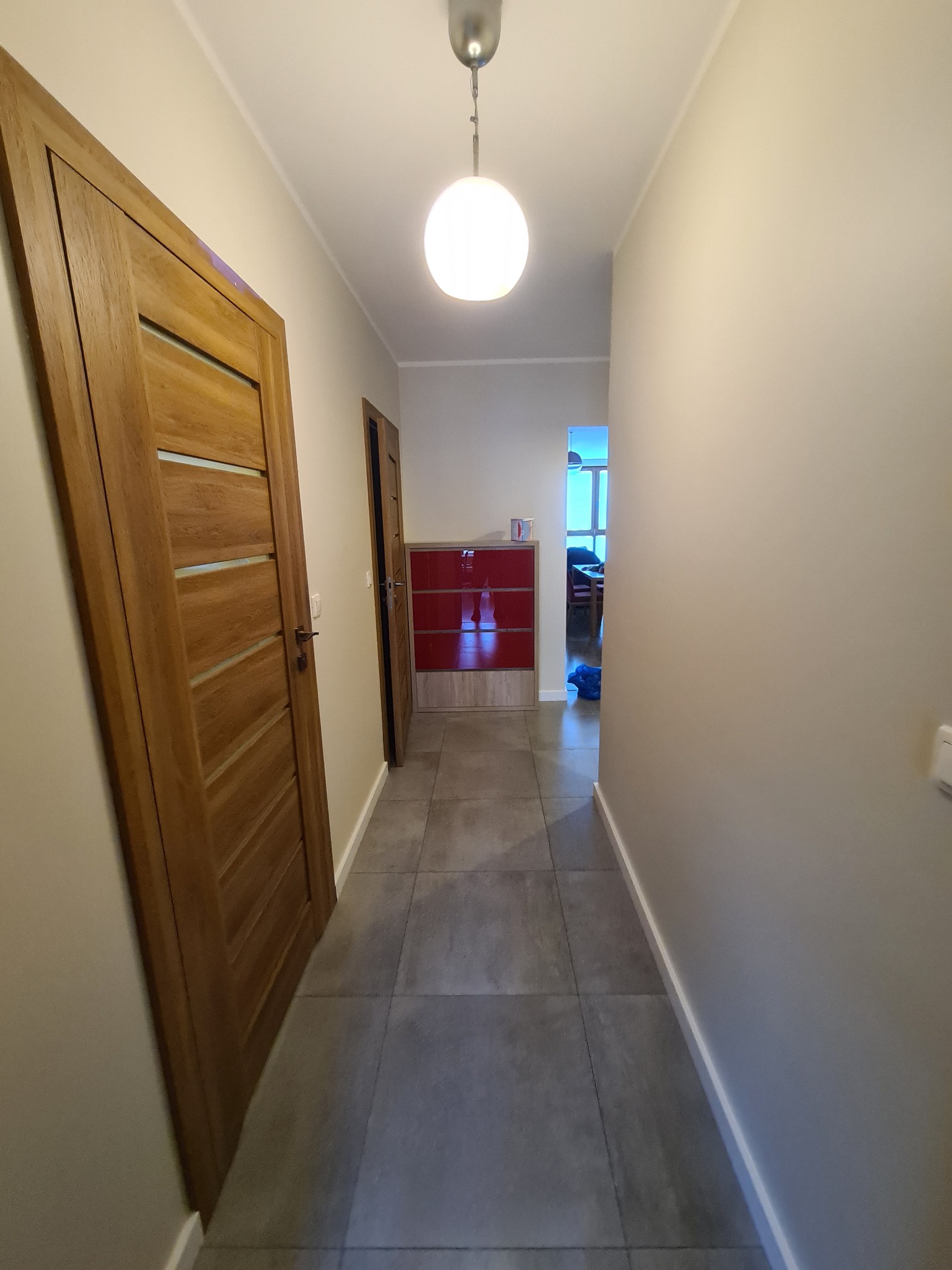 Korytarz z szarymi płytkami podłogowymi, beżowymi ścianami, drewnianymi drzwiami z poziomymi przeszkleniami i lampą sufitową w kształcie kuli.