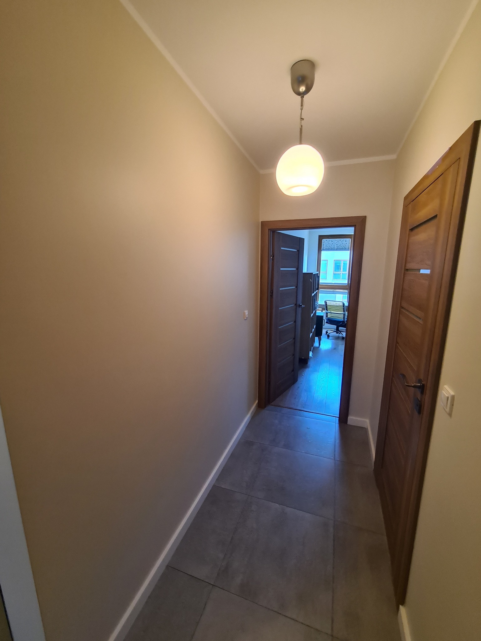 Korytarz w mieszkaniu po remoncie, ściany pomalowane na beżowo, szara podłoga z płytek, widoczne dwie drewniane ościeżnice z drzwiami, w głębi widok na biuro z oknem.