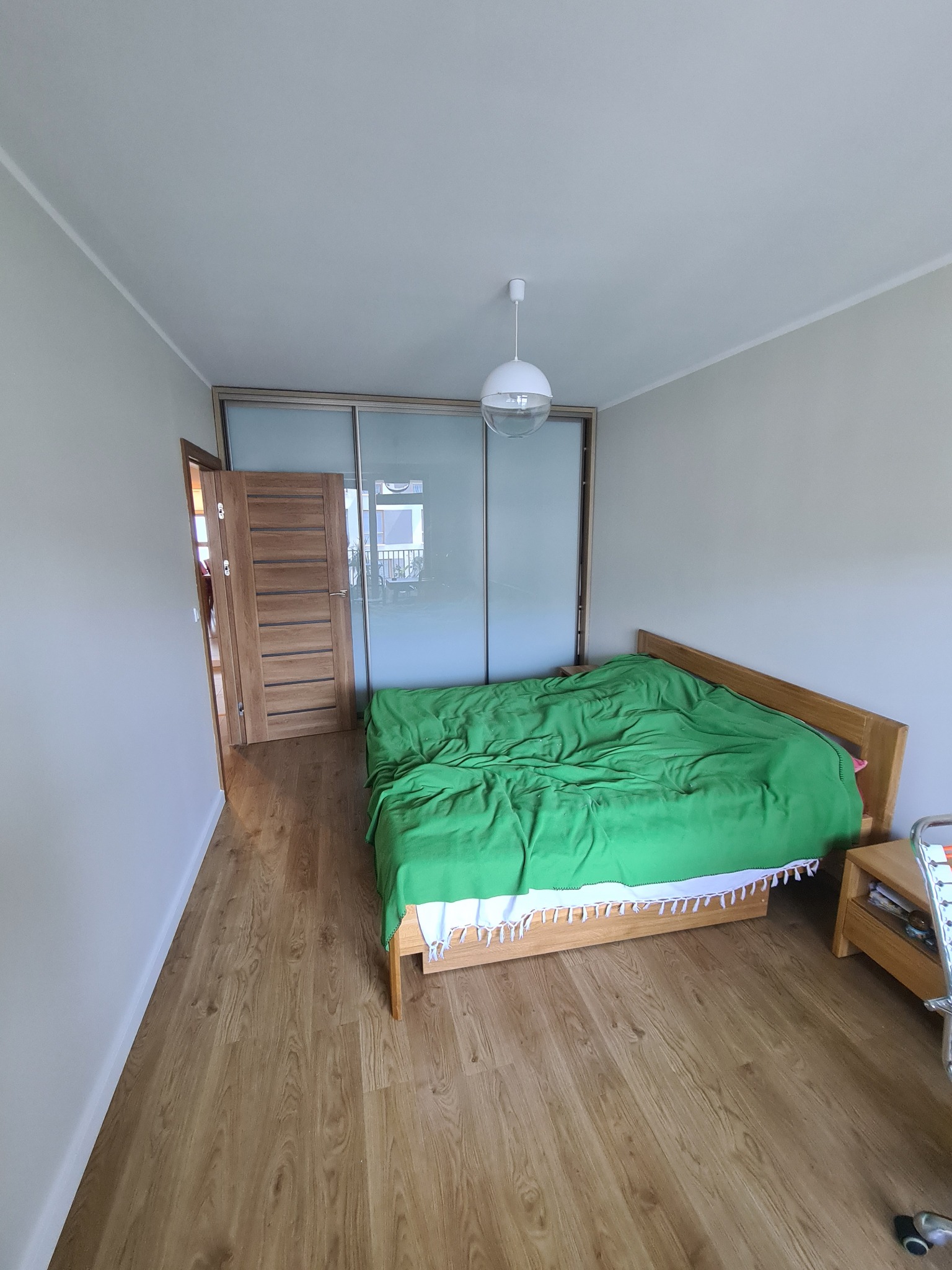 Jasne wnętrze sypialni z drewnianą podłogą i łóżkiem z zieloną narzutą, widok na przesuwne drzwi szafy wnękowej i otwarte drzwi do innego pomieszczenia, ściany pomalowane na jasny odcień szarości.