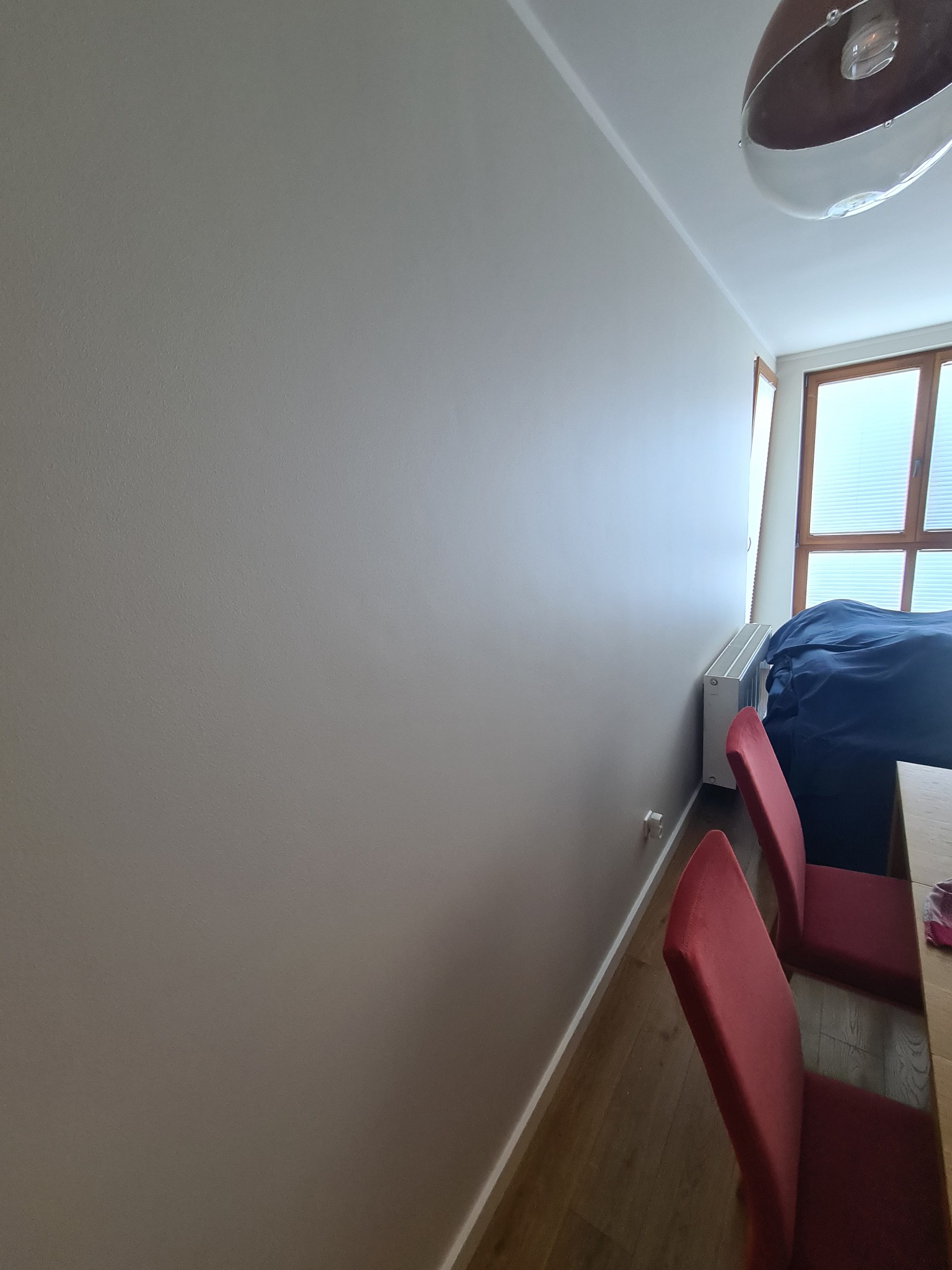 Świeżo pomalowana ściana w odcieniach szarości w jasnym pokoju z dwoma czerwonymi krzesłami, widoczny fragment drewnianej podłogi i grzejnik pod oknem.
