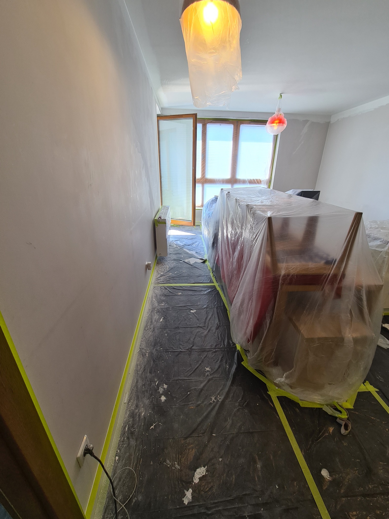 Przygotowanie pokoju do prac wykończeniowych: ściana po szpachlowaniu, podłoga i meble zabezpieczone folią i taśmą malarską, wisząca żarówka osłonięta folią.
