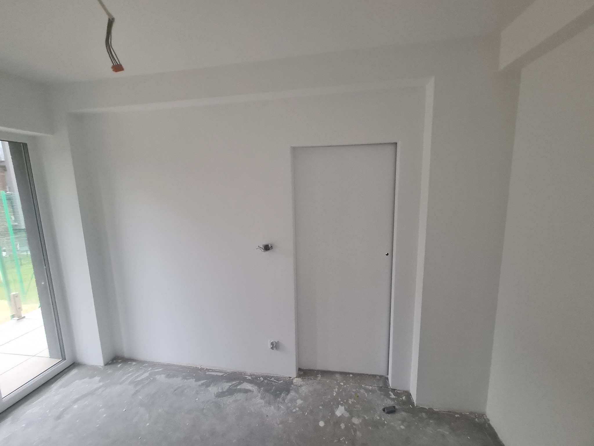 Wnętrze po świeżym malowaniu na biało, widoczne gniazdka elektryczne i kable wystające z sufitu, podłoga z plamami farby.