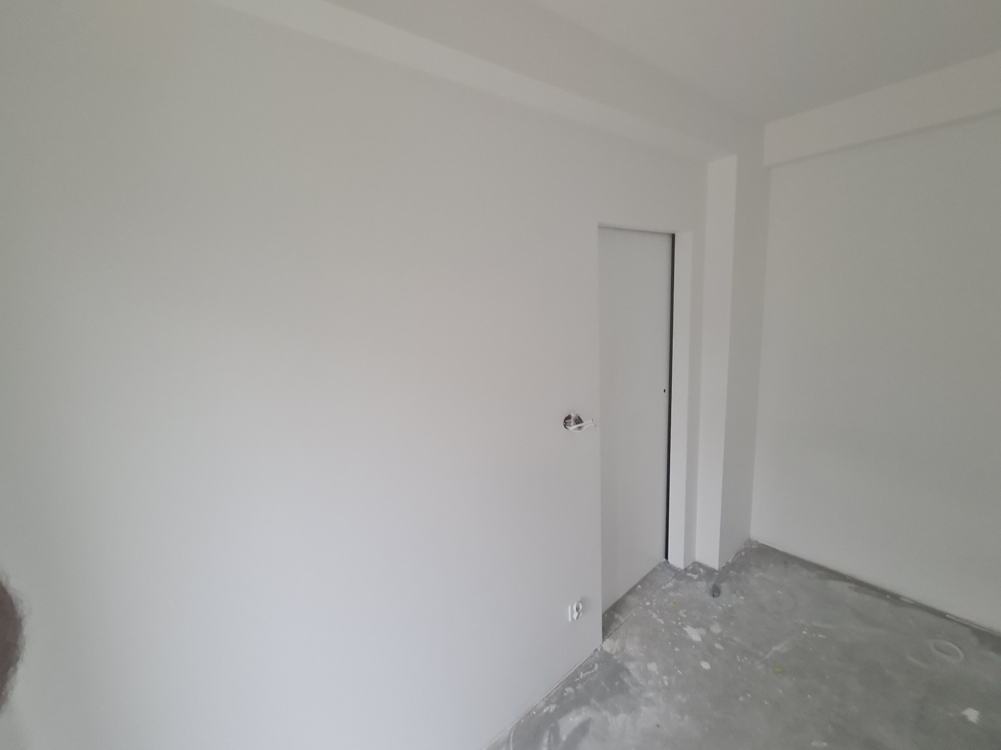 Wnętrze pokoju po świeżym pomalowaniu na biało, widoczne ślady farby na szarej podłodze, białe drzwi i puszka elektryczna bez osprzętu na ścianie.