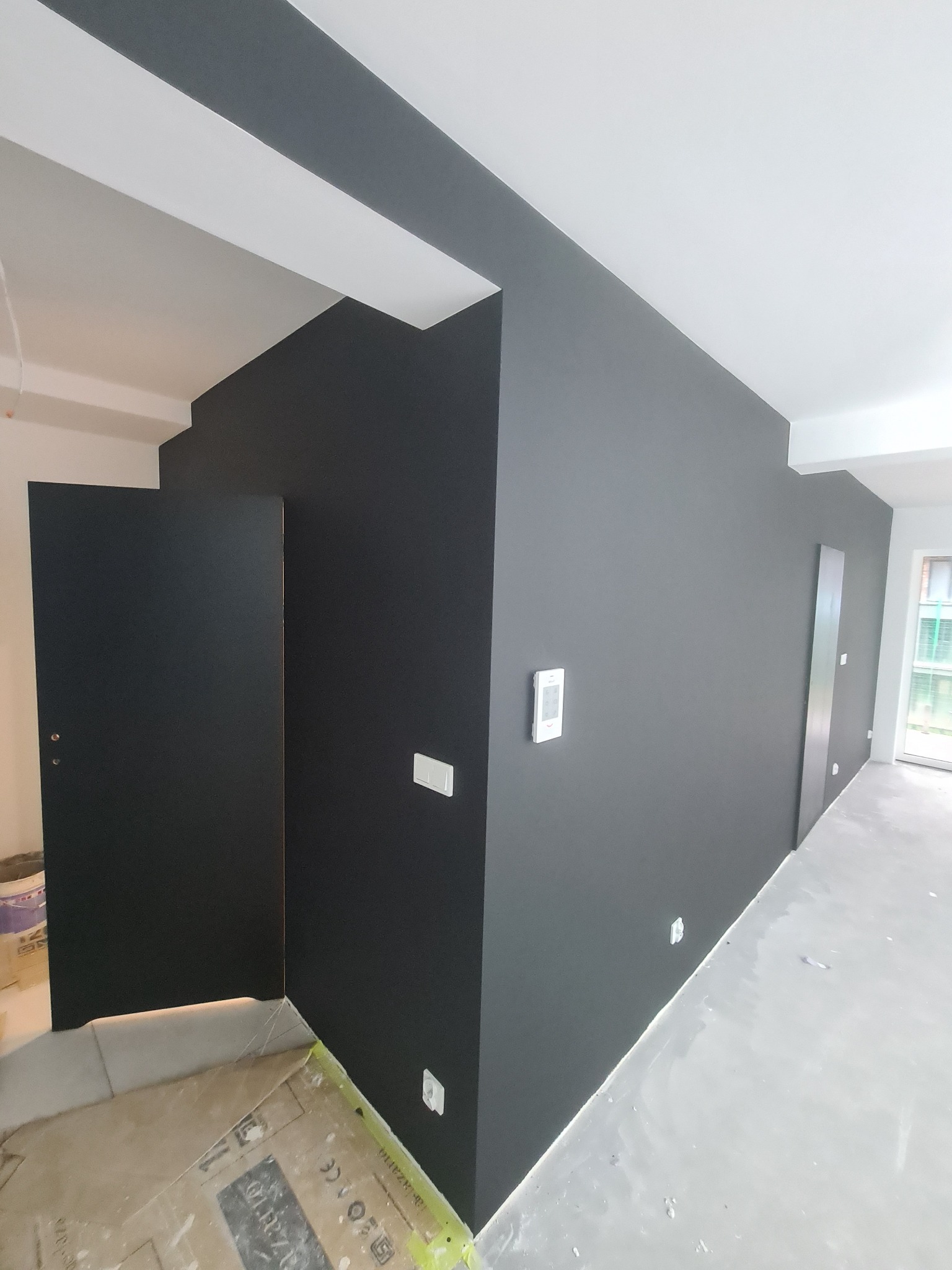 Wnętrze po malowaniu ścian na ciemny odcień szarości, widoczna ościeżnica drzwiowa, białe listwy przypodłogowe i zabezpieczona podłoga przed zabrudzeniem podczas prac malarskich.