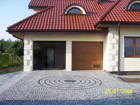 Podjazd do garażu wyłożony szarą kostką z dekoracyjnym wzorem koncentrycznych okręgów z ciemniejszej kostki, widok na budynek z dwoma bramami garażowymi i czerwoną dachówką.